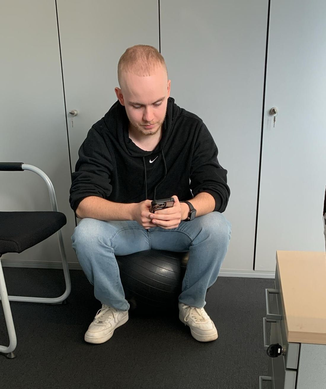 Yannick früher im Büro: Er sitzt auf einem Sitzball vor grauen Schränken und schaut aufs Handy.